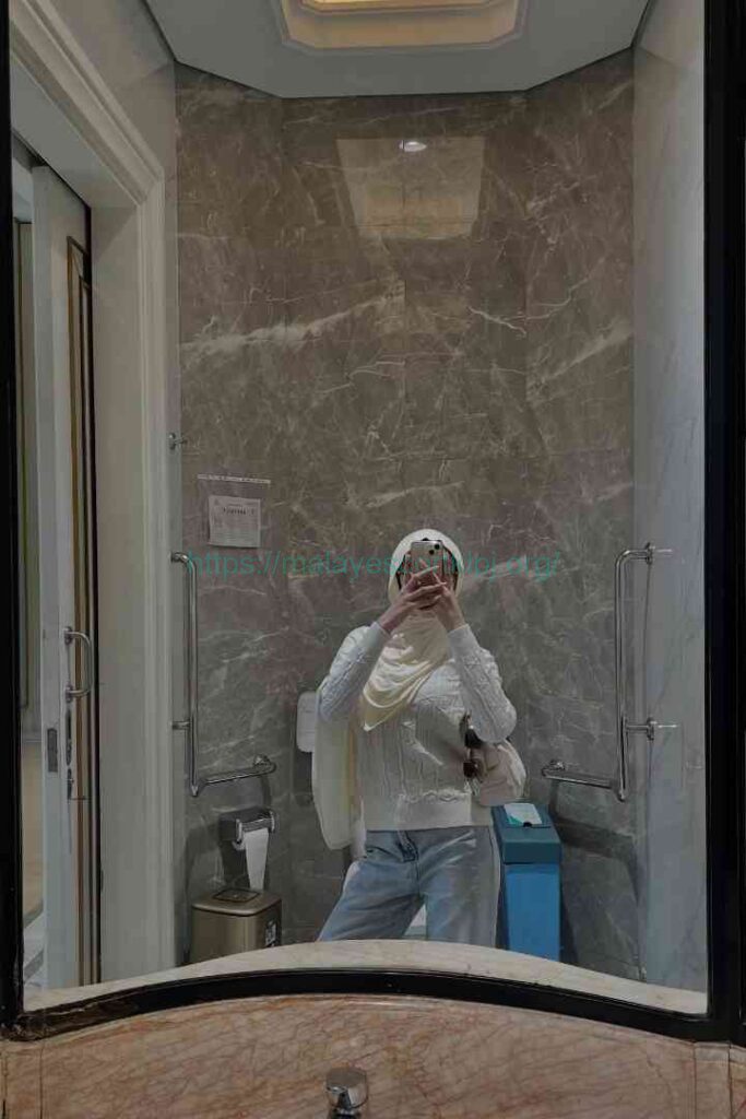 Kesya in a white sweater and hijab takes a mirror selfie in a modern bathroom